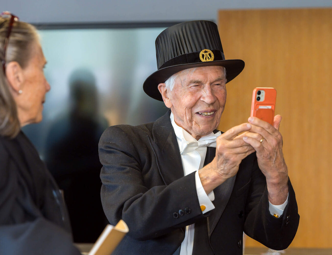 Elderly person in formal black suit and top hat takes a photo with an orange smartphone indoors