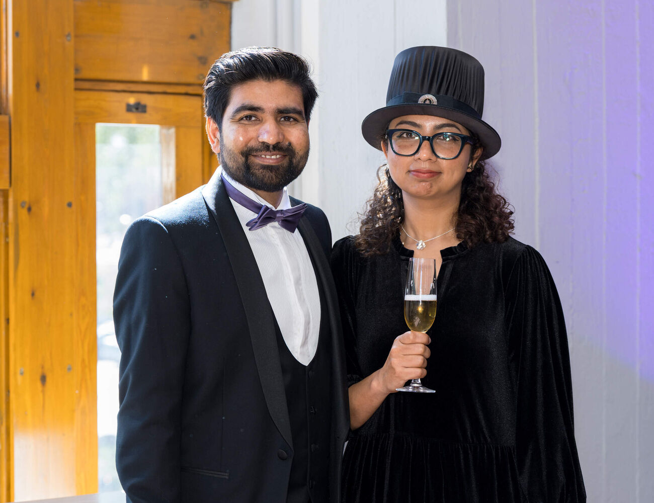Two formally dressed people indoors; one in a suit, one in a black dress and top hat holding a champagne glass