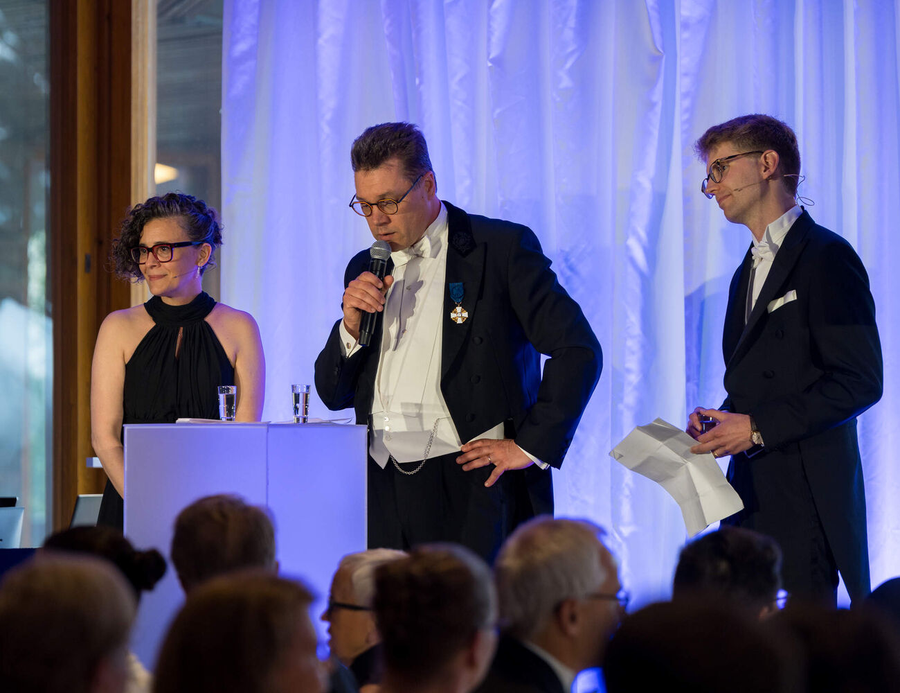 Three speakers in formal black and white evening wear on stage at a blue-lit event