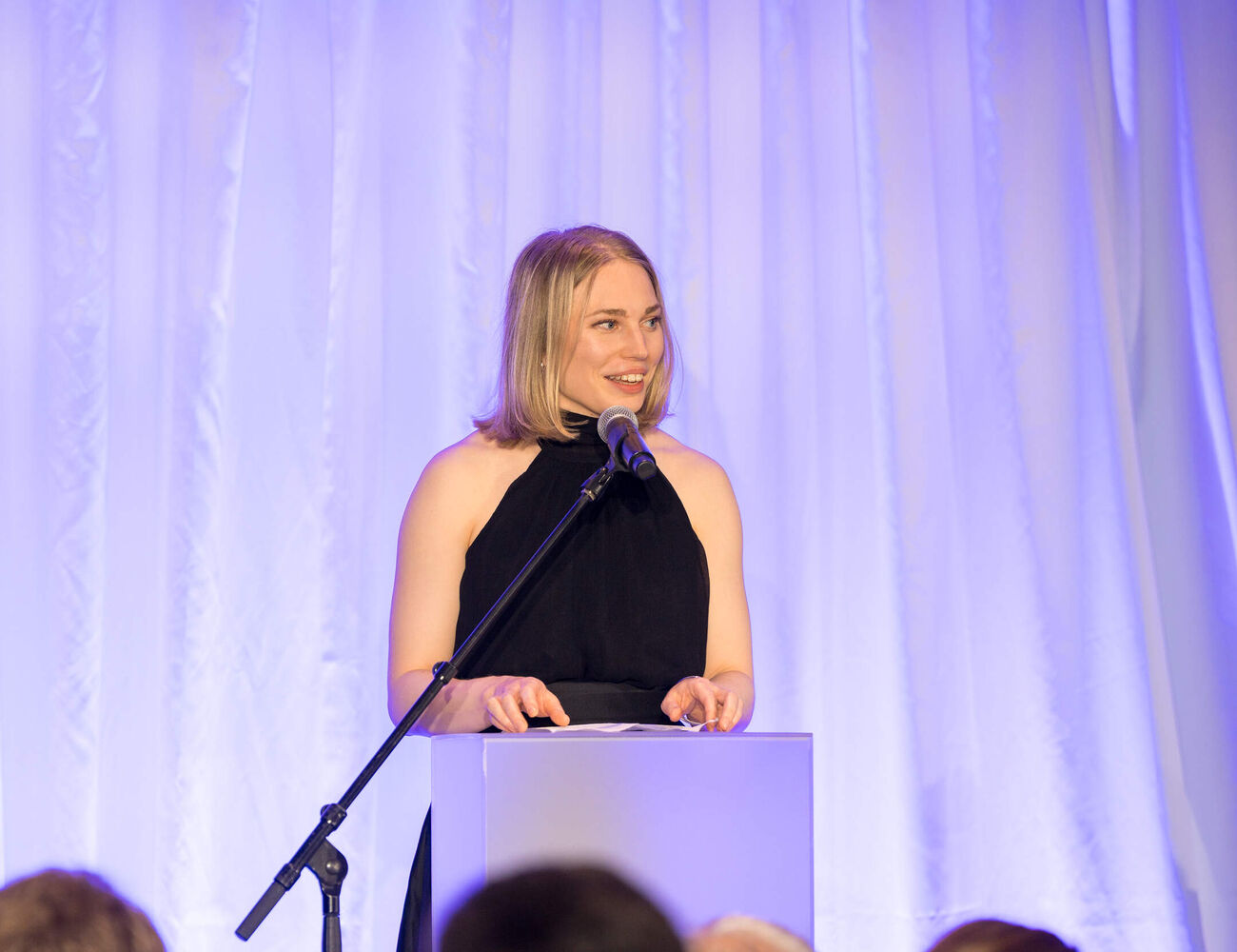 Speaker in a black dress at a podium, addressing an audience against purple-lit curtains