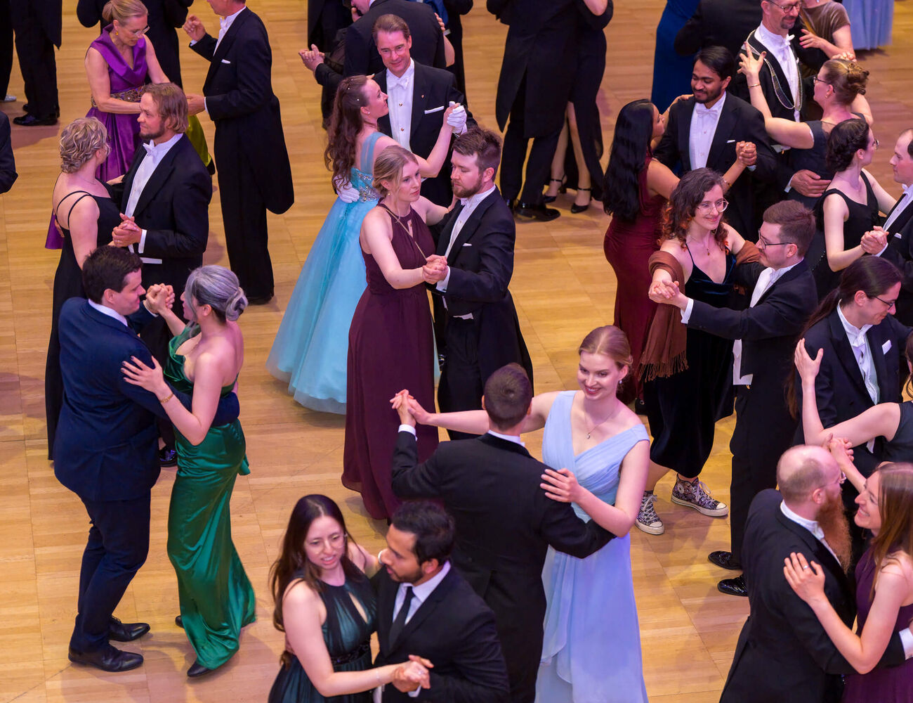 Many couples in formal suits and colourful gowns waltz together on a polished wooden ballroom floor