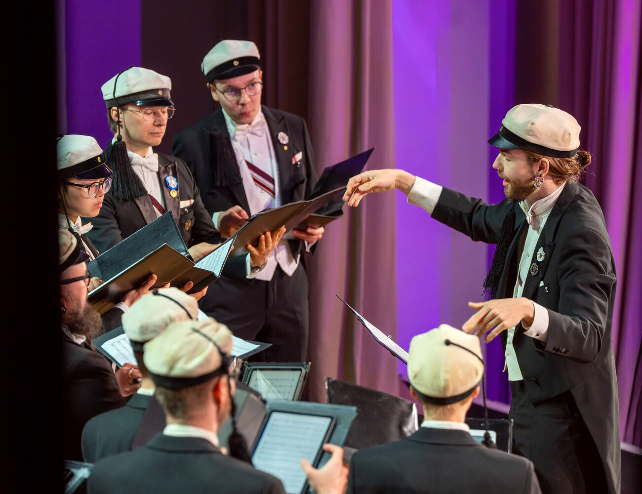 Choir in dark suits and white caps sings on stage as a conductor leads under purple light