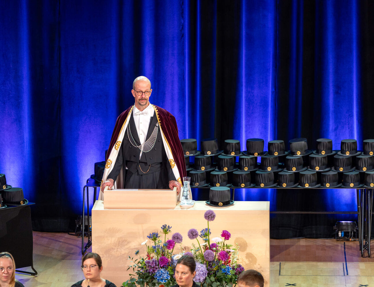 Robed speaker at a podium on stage with doctoral hats and flowers, before blue curtains