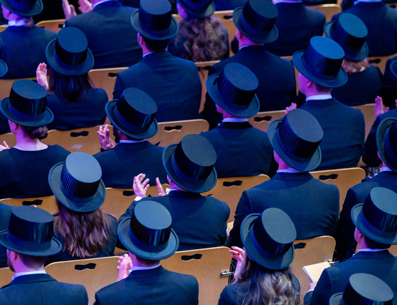 Audience in dark suits and black top hats seated in rows of wooden chairs at a formal ceremony