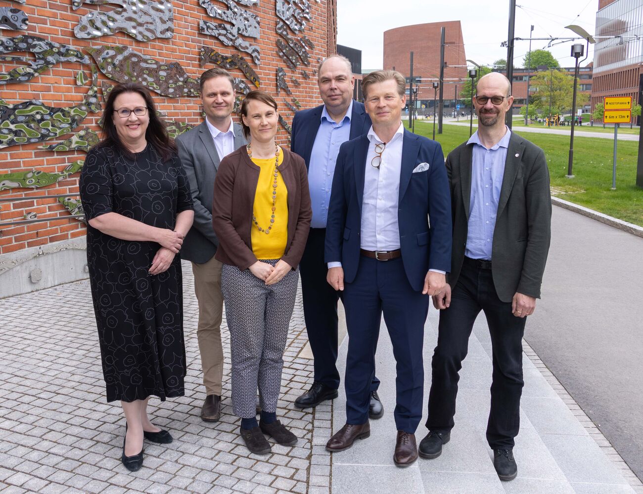 Six adults in smart clothing pose outside a brick building with wall art and a modern campus in the background
