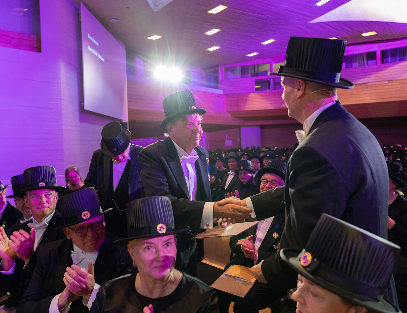 Formal ceremony in purple-lit hall, two people in black tailcoats and top hats shaking hands