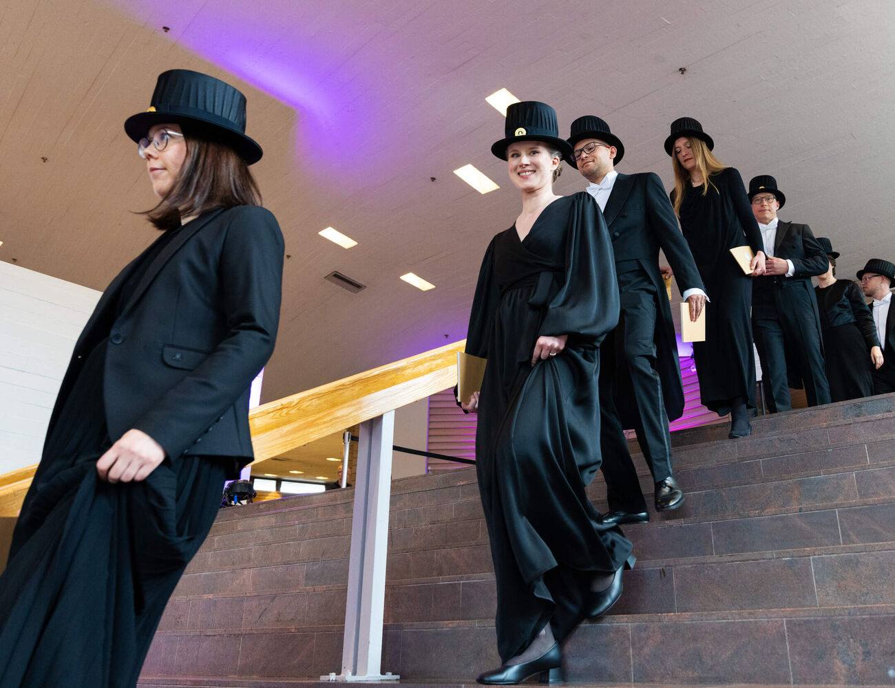People in black formal dress and top hats walk down indoor stairs holding light-coloured books