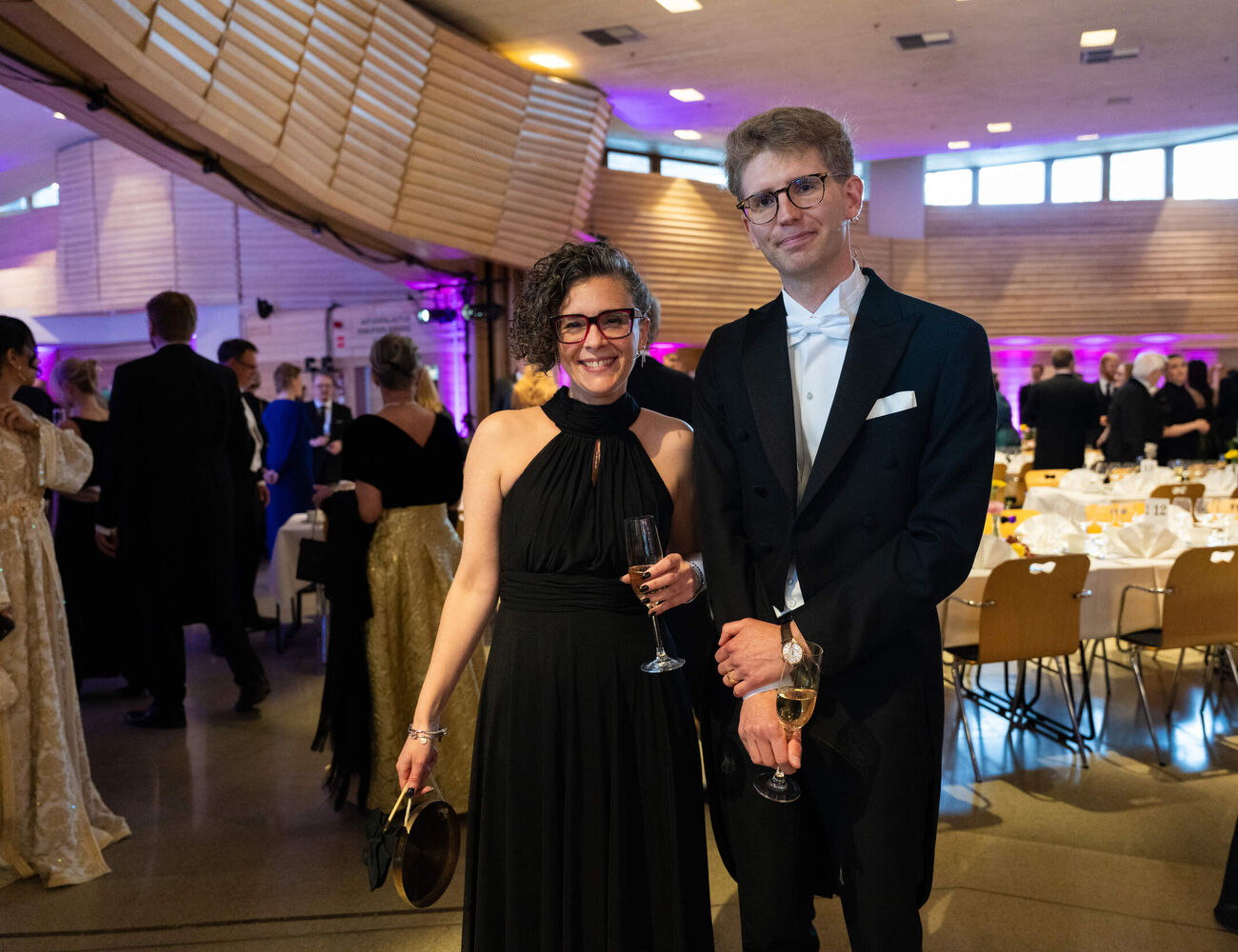 Well-dressed couple holding champagne glasses at a formal indoor gala with round tables and guests in evening wear