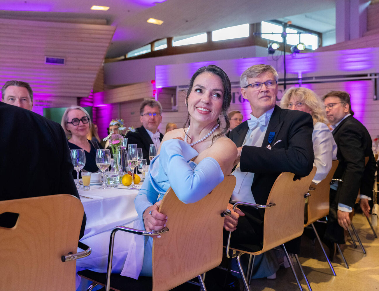 Formal dinner guests seated at round tables with drinks in a hall lit by pink and purple lights