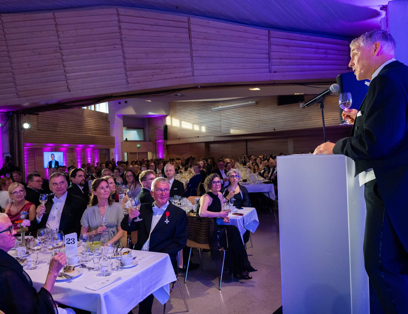 Speaker in black tailcoat giving a toast to a seated banquet audience in a purple-lit wooden hall