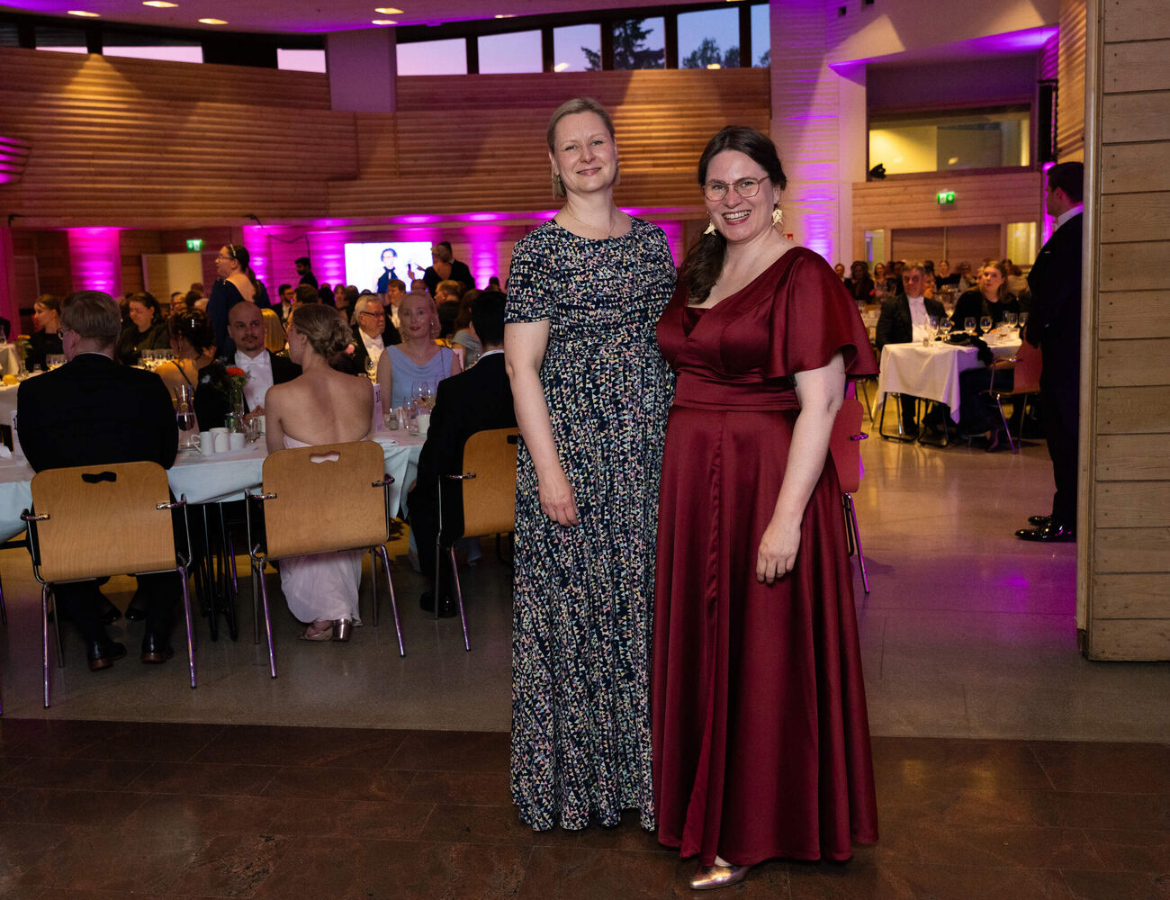 Two women in long dresses pose at a formal dinner, guests seated at tables in a purple-lit hall