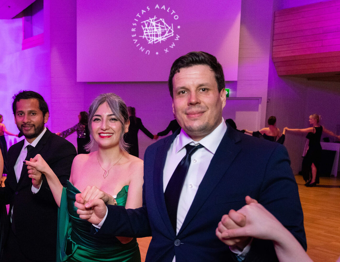 People in formal dress dance in a ballroom with purple lighting and an Aalto University logo on the wall.