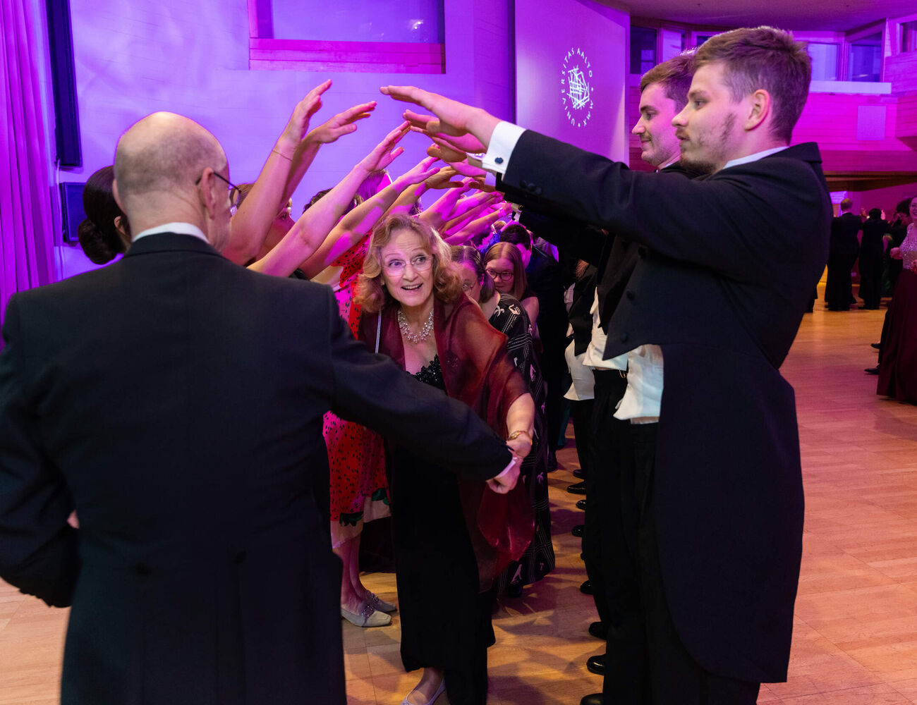 People in formal wear form an arm tunnel for dancers at a purple-lit ballroom event