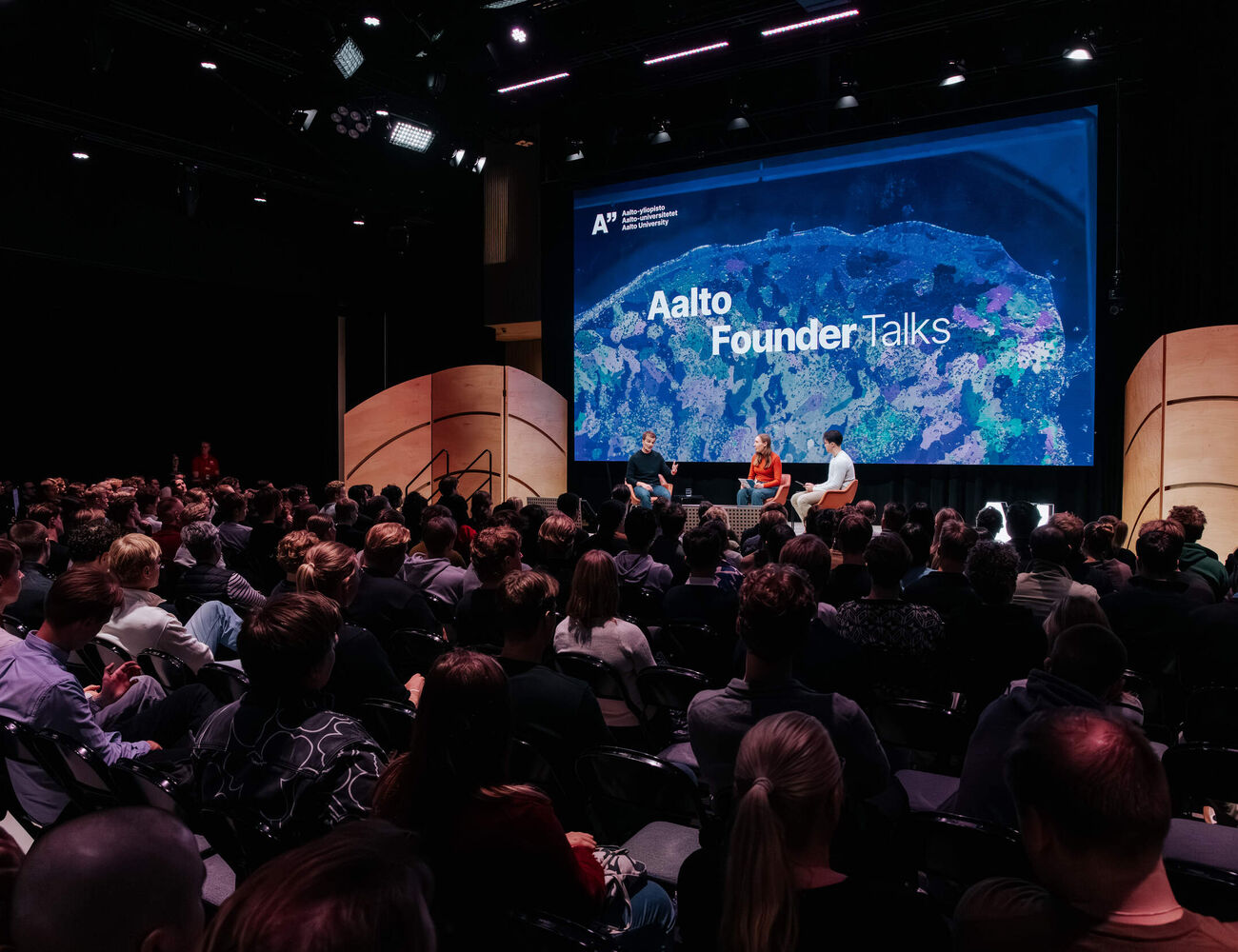 Audience watching three speakers on stage at Aalto Founder Talks in a dark auditorium with a large blue screen.