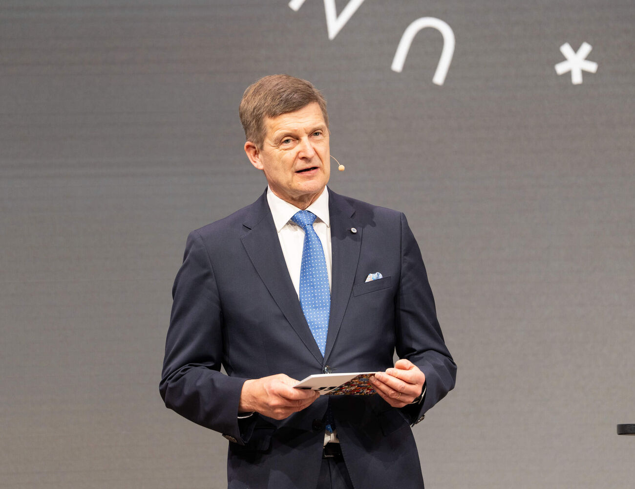 President Ilkka Niemelä in dark suit and blue tie speaks on stage, holding notes, with symbols on grey backdrop.