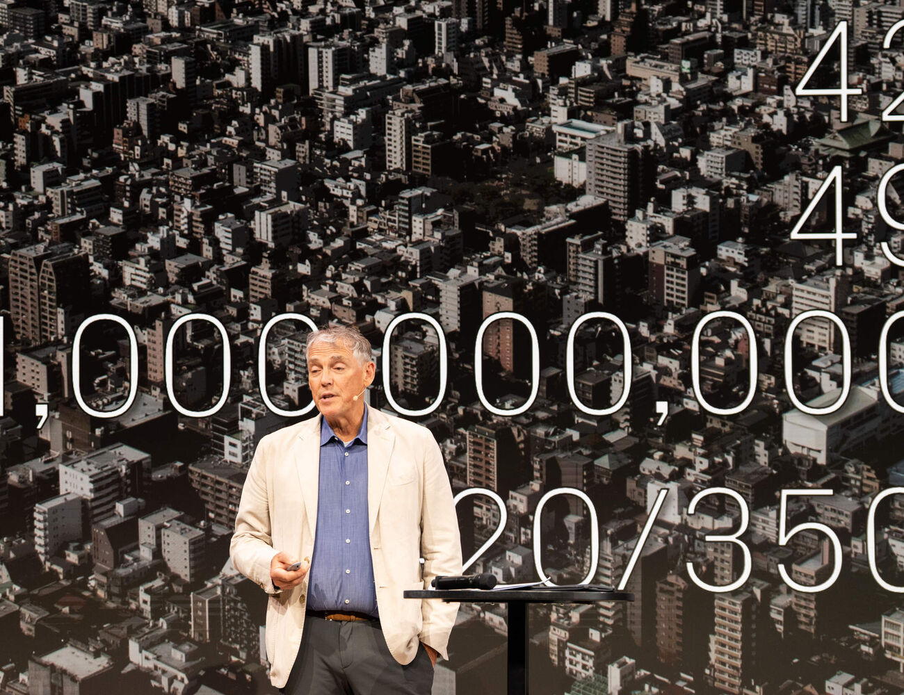 Alan Organschi in beige jacket on stage, city aerial backdrop with large white numbers