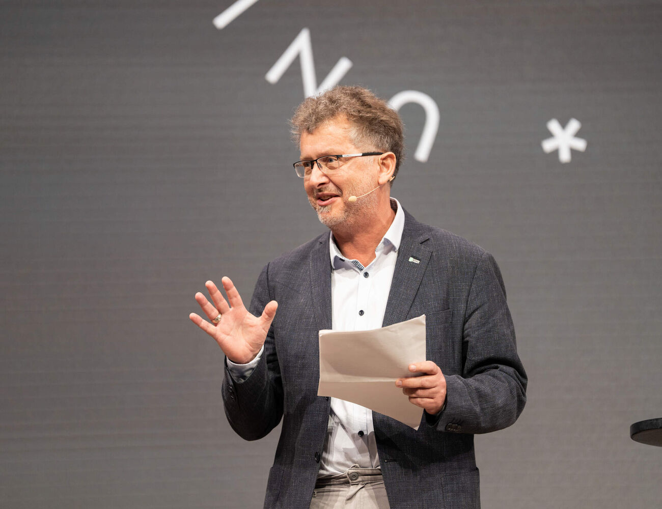 Person in grey blazer speaking on stage, holding papers, with abstract white symbols on grey background