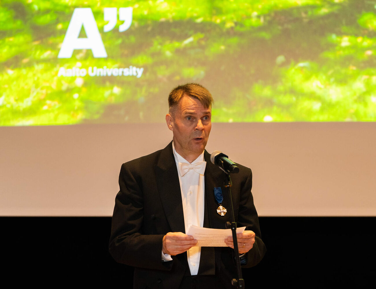 Person in black tailcoat speaking at a microphone, Aalto University logo on bright green screen behind