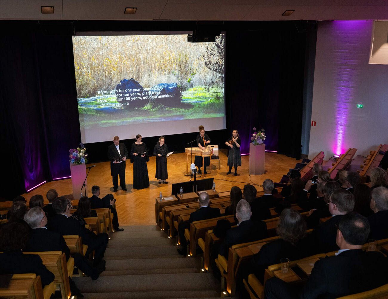 Audience in a modern hall watching four speakers on stage under a large nature image with a proverb.