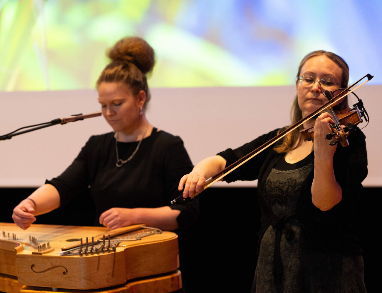 Two women in black perform on stage, one on a wooden zither, the other playing a violin