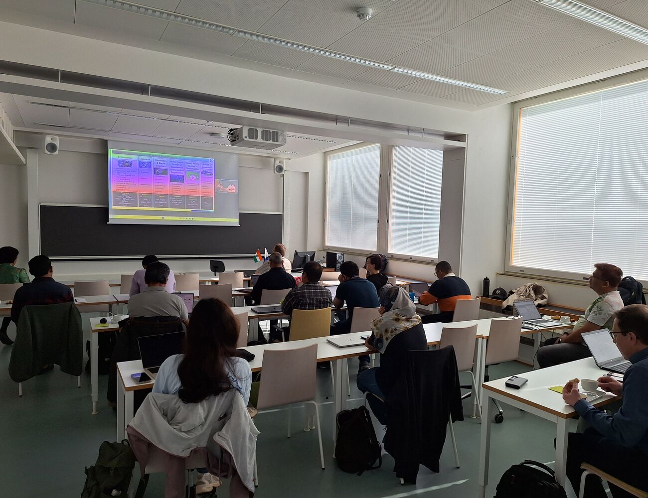 Students sit at desks with laptops in a bright classroom, watching a colourful slide on a projector screen.