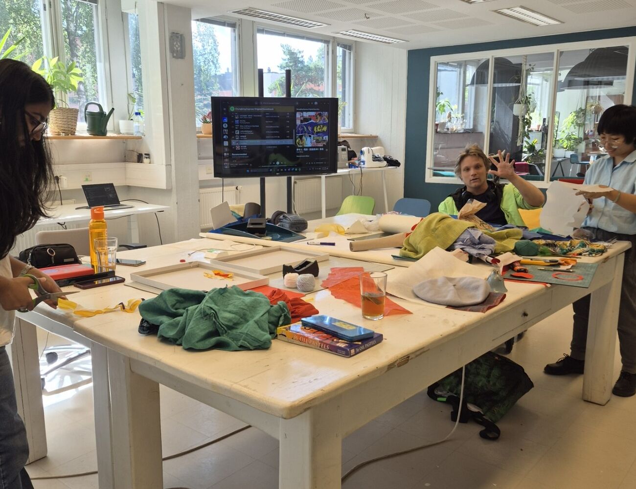 Three people crafting at a large white table covered with colourful fabrics and art supplies in a bright studio