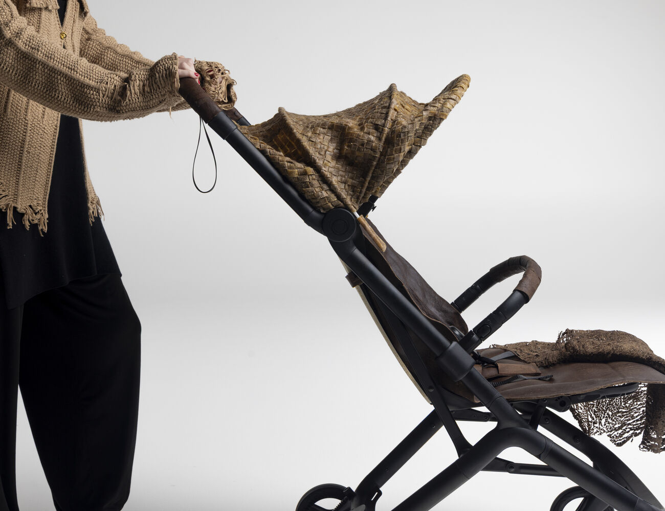 Person in loose beige jumper pushing an empty brown stroller with textured canopy and blanket