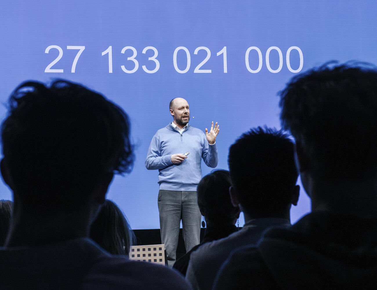 Mies esitelmöi lavalla yleisön edessä, takana sininen näyttö ja luku 27 133 021 000
