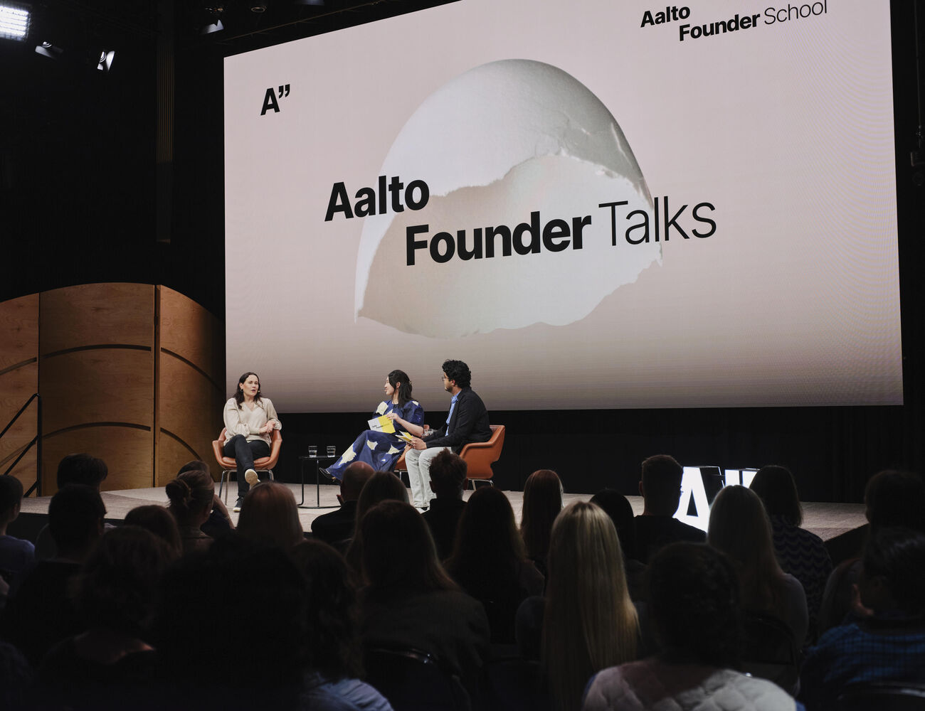 Kolme puhujaa Aalto Founder Talks -lavalla oransseilla tuoleilla, valkoinen tausta ja yleisö edessä.