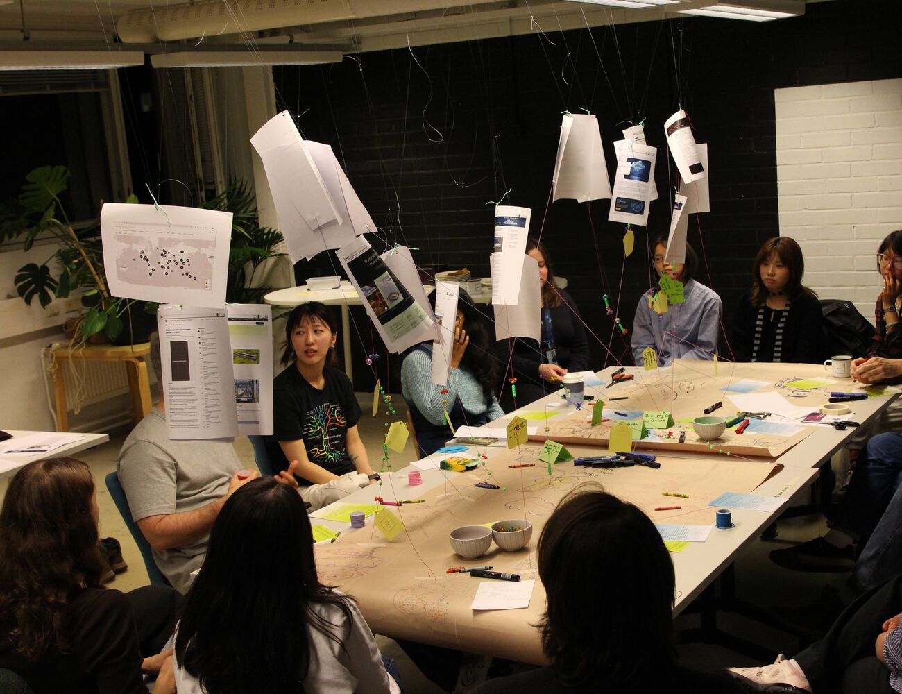 People sit round a table covered in notes and string, with papers hanging above during a creative workshop.