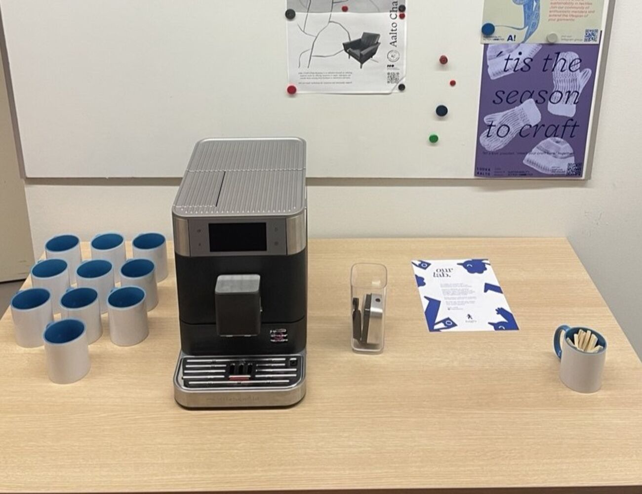 Coffee machine on wooden table with mugs, stirrers, flyers and whiteboard with posters behind.