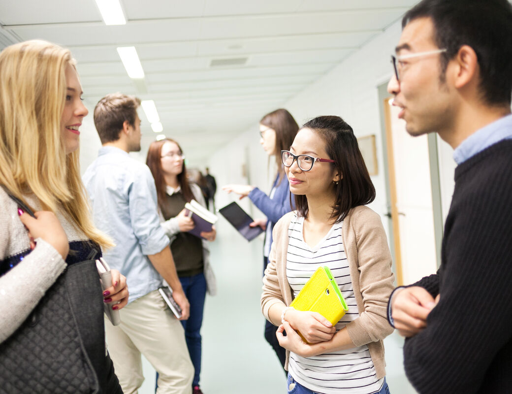 Several students, women and men discussing in the school hallway. One of the women is holding a yellow book.