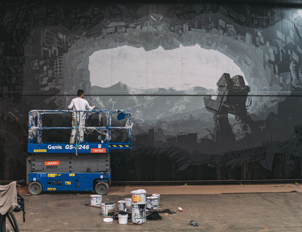 Artist Jesse stands on a scissor lift to paint the greyscale mural in Keran Hallit