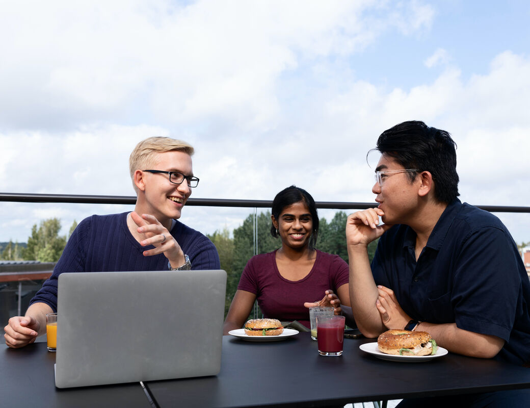 Three students eating a snack and discussing over a computer on a rooftop terrace. 