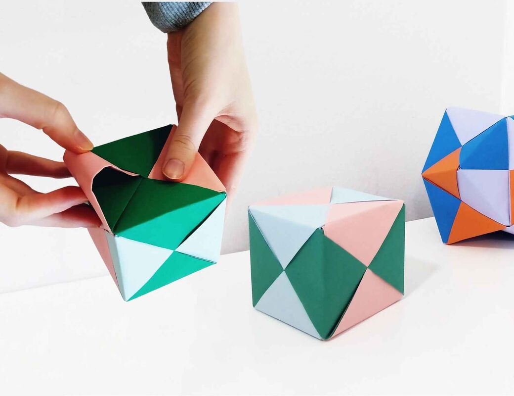 Kädet taittelevat värikästä origamikuuutiota, taustalla origamikuutio sekä tähtimäistetty oktaedri