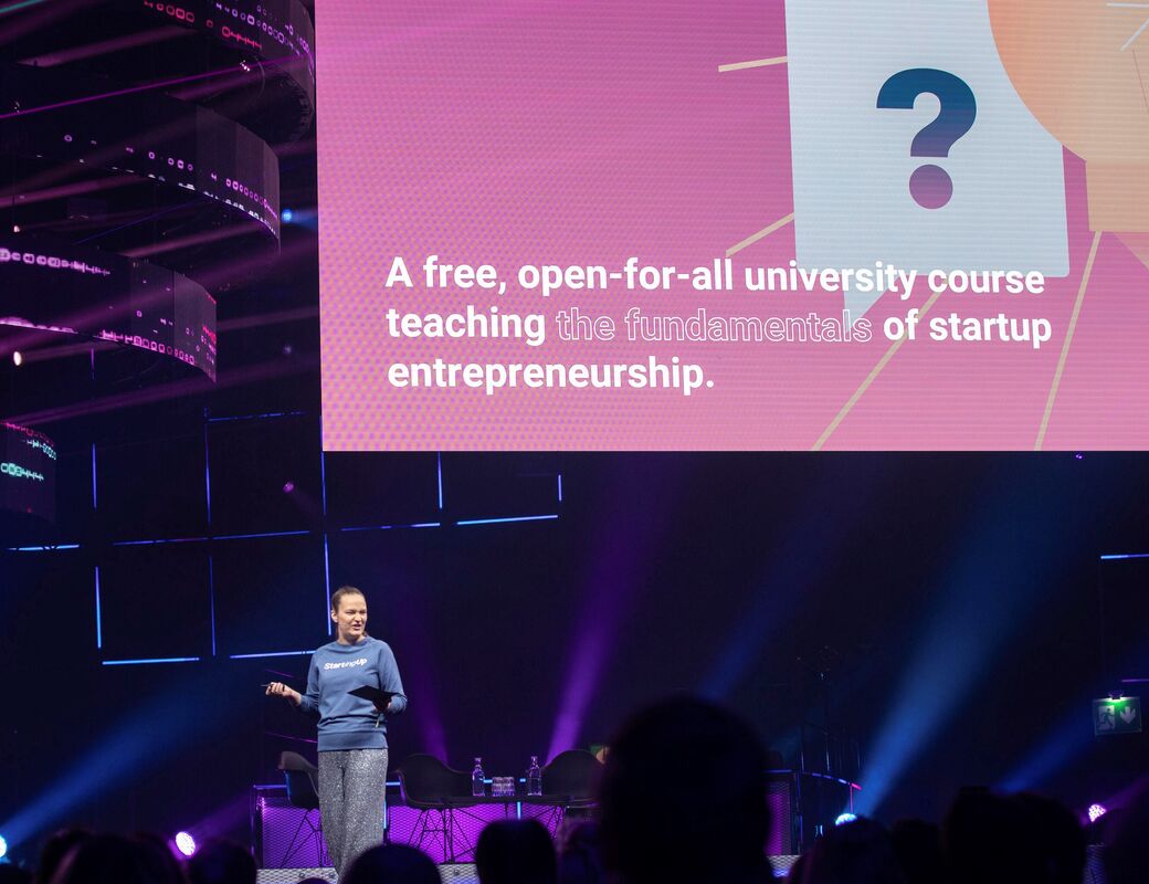A woman stands on stage at Slush, presenting Starting Up online course, lights are purple