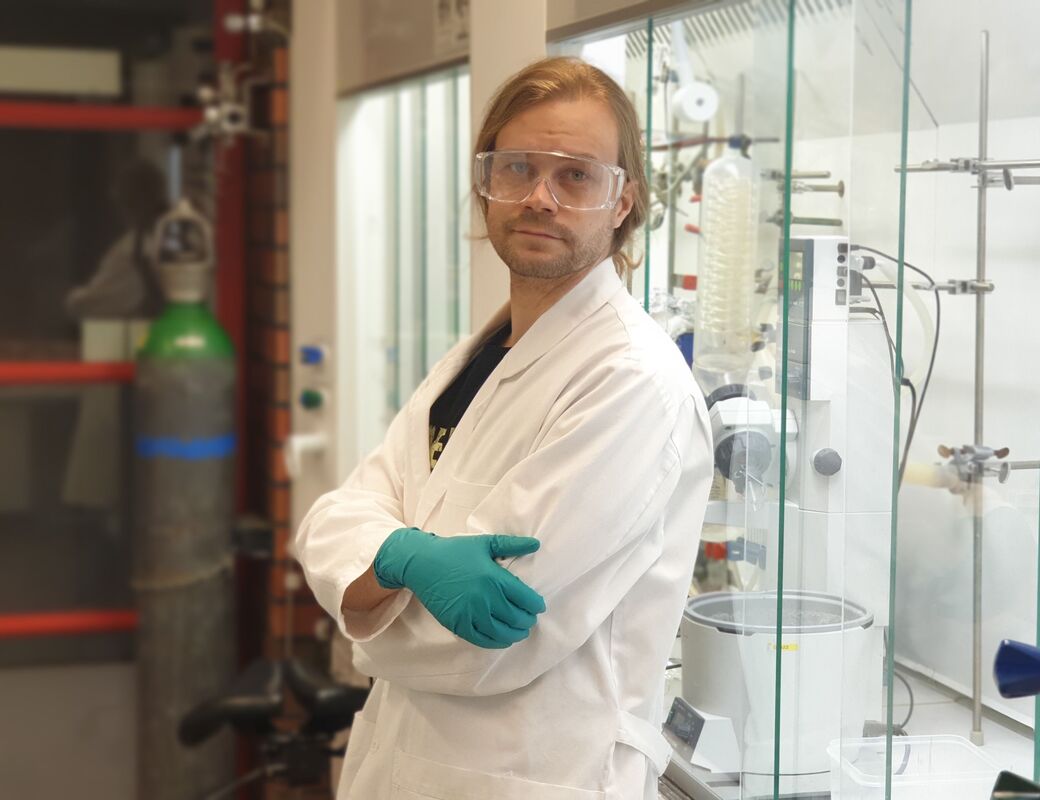 A person in a lab coat and green gloves stands in a science laboratory with various equipment and machinery.