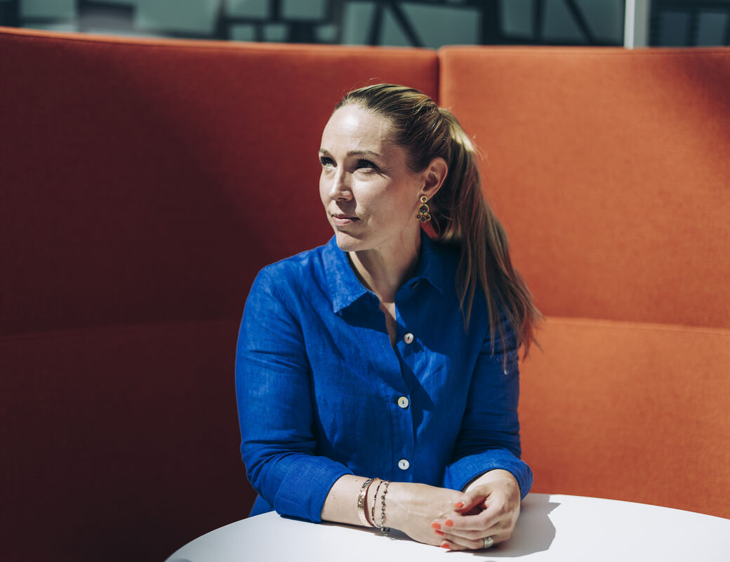Siniseen asun pukeutunut Maija Jokela istumassa pyöreän pöydän ääressä neuvotteluhuoneessa, taustalla suuren sohvan oranssi selkänoja.