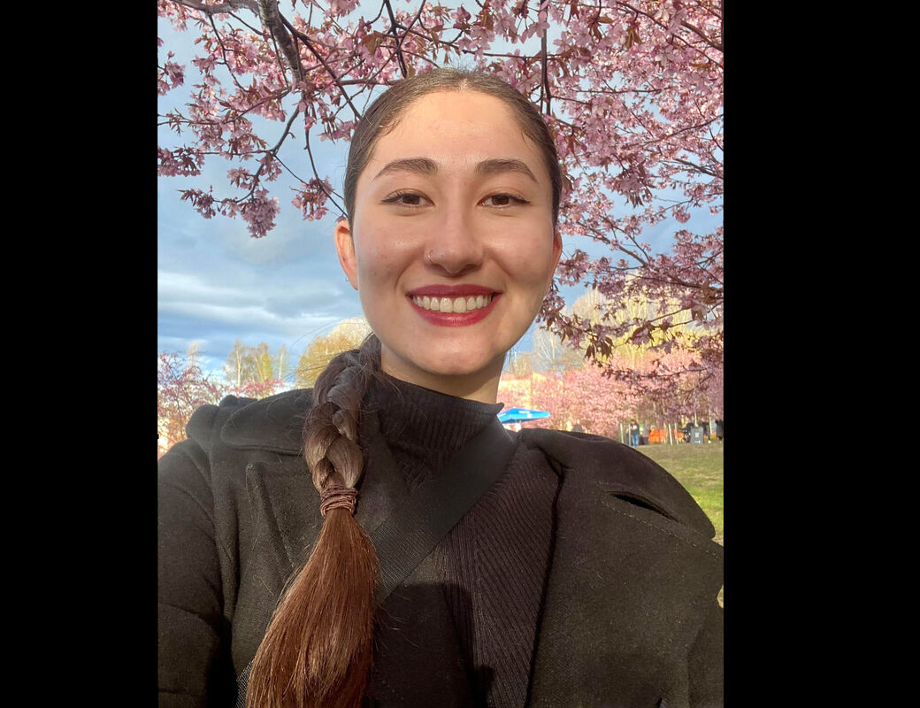 Person with braided hair, wearing a black jacket under cherry blossom trees.