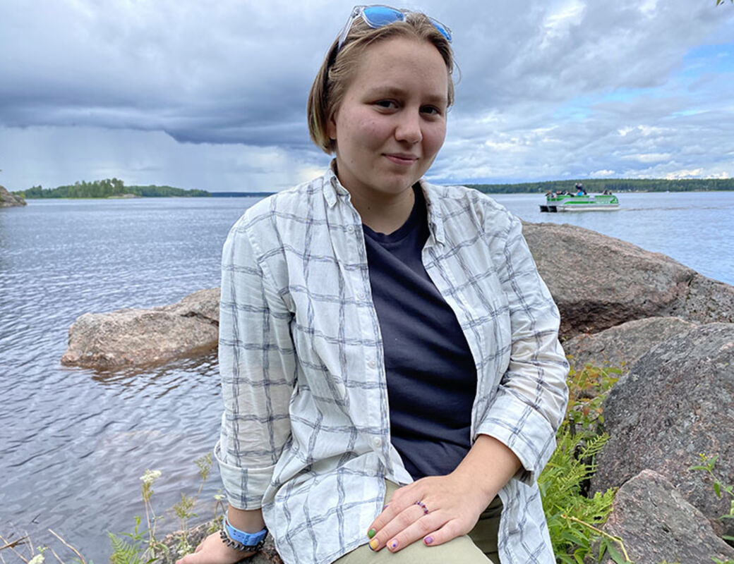 Person sitter på stenar vid en sjö, med en rutig skjorta och mörk topp. En båt är i bakgrunden.