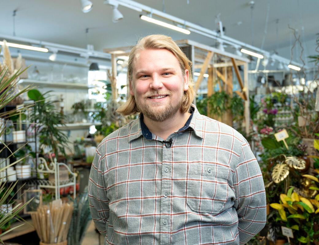 A person wearing a grey checked shirt is standing in a store filled with various plants and gardening supplies.
