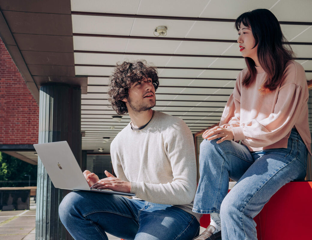 Two students sitting and discussing outside while one student is holding a laptop in his knee while listening to the other student.