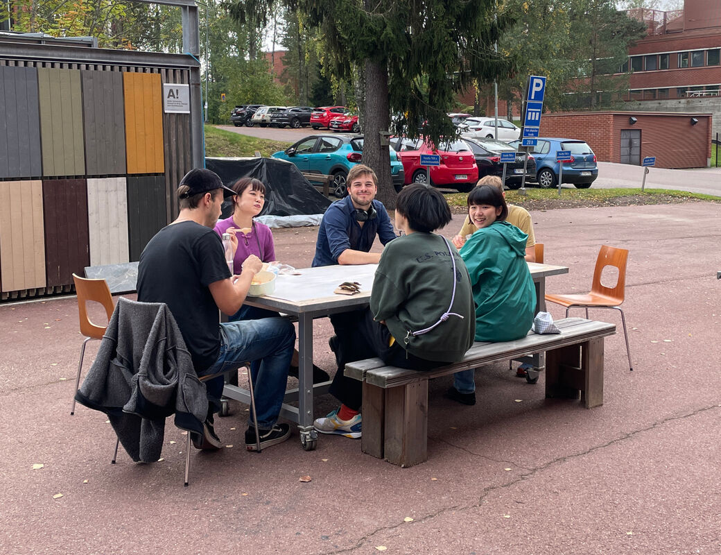 Six students sitting around a table outside enjoying the weather and smiling at camera