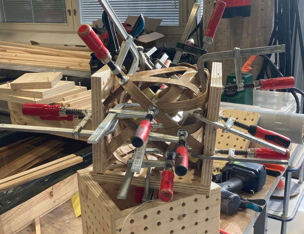 Cube made of bent strips of oak, still in the square plywood mold with big clamps holding it while glue dries