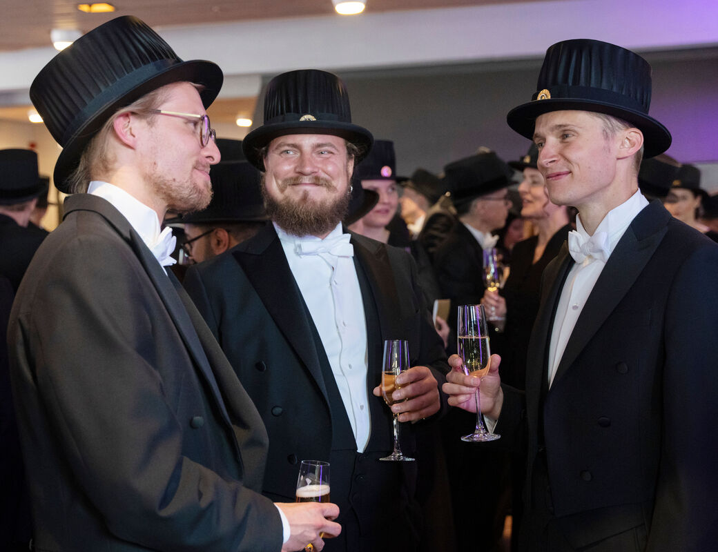 Kolme juhlapukuista miestä hymyilee kuohuviinilasit käsissään