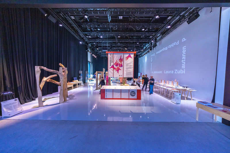 An exhibition hall with art and design displays, people viewing various installations. Black curtains and bright floor lighting.