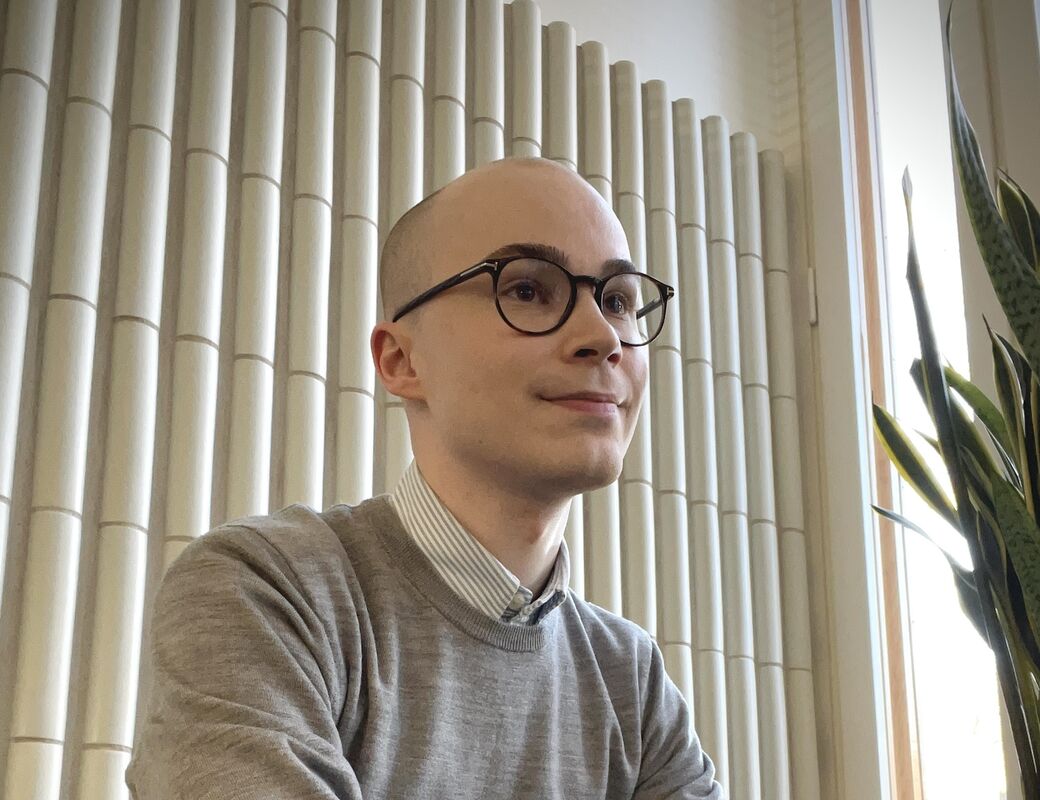 A PhD researcher with dark glasses sits near a window on the Aalto University campus.