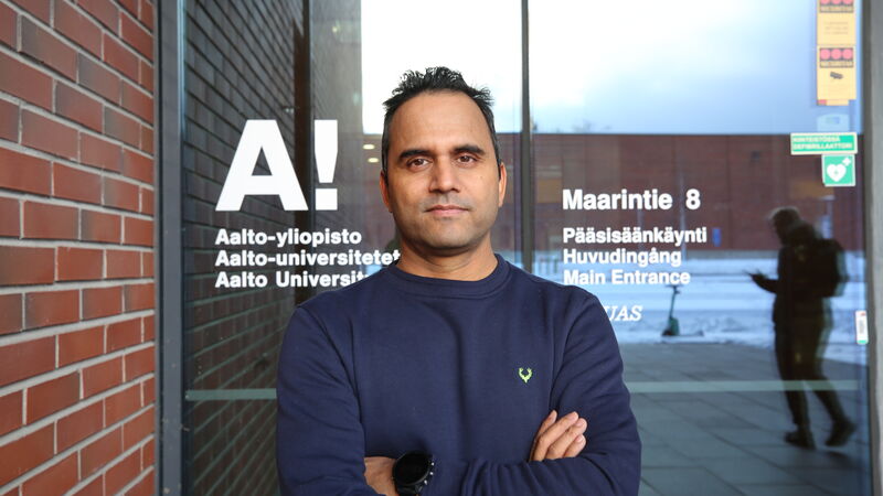 Person with crossed arms standing in front of Aalto University entrance. Brick wall and glass door with text behind.