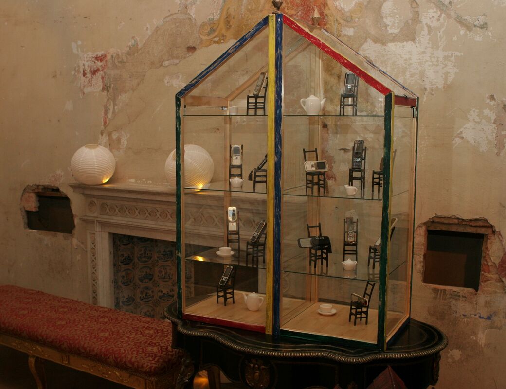 Glass display case shaped like a house with mobile phones on small chairs. Background shows an old wall and a fireplace.