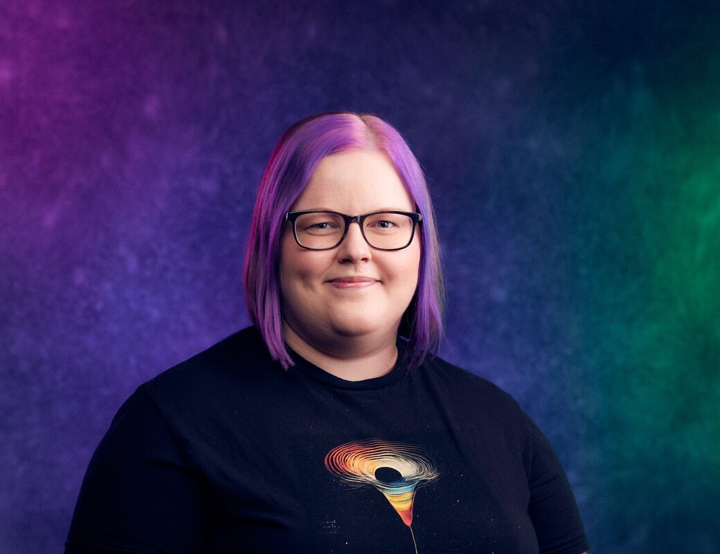 Henkilö, jolla on olkapäille ulottuvat hiukset ja musta paita, jossa on värikäs abstrakti kuvio. Tausta on liukuvärjätty violetti, sininen ja vihreä.